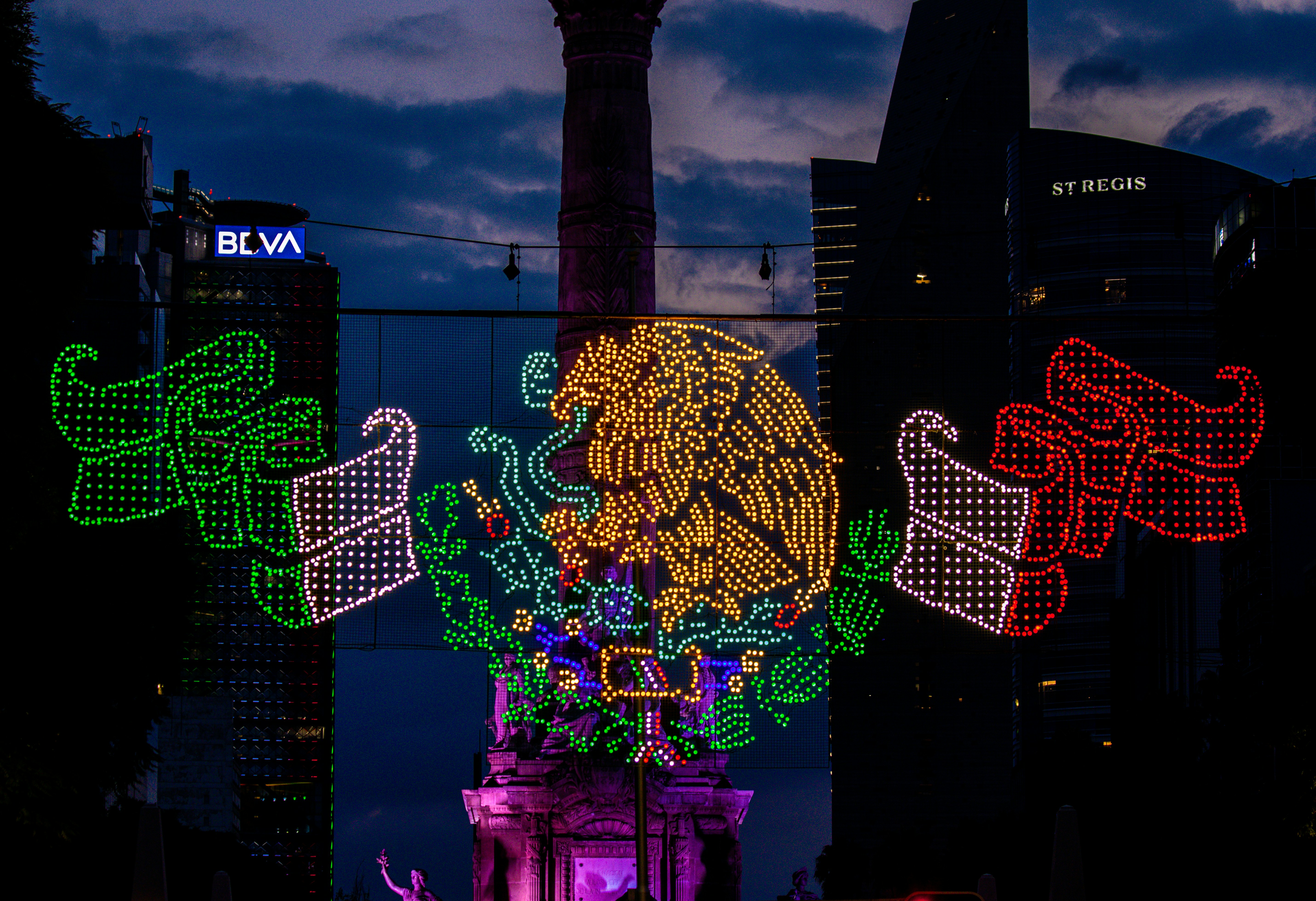 An illuminated light display of the Mexican coat of arms above Paseo de la Reforma in Mexico City at dusk