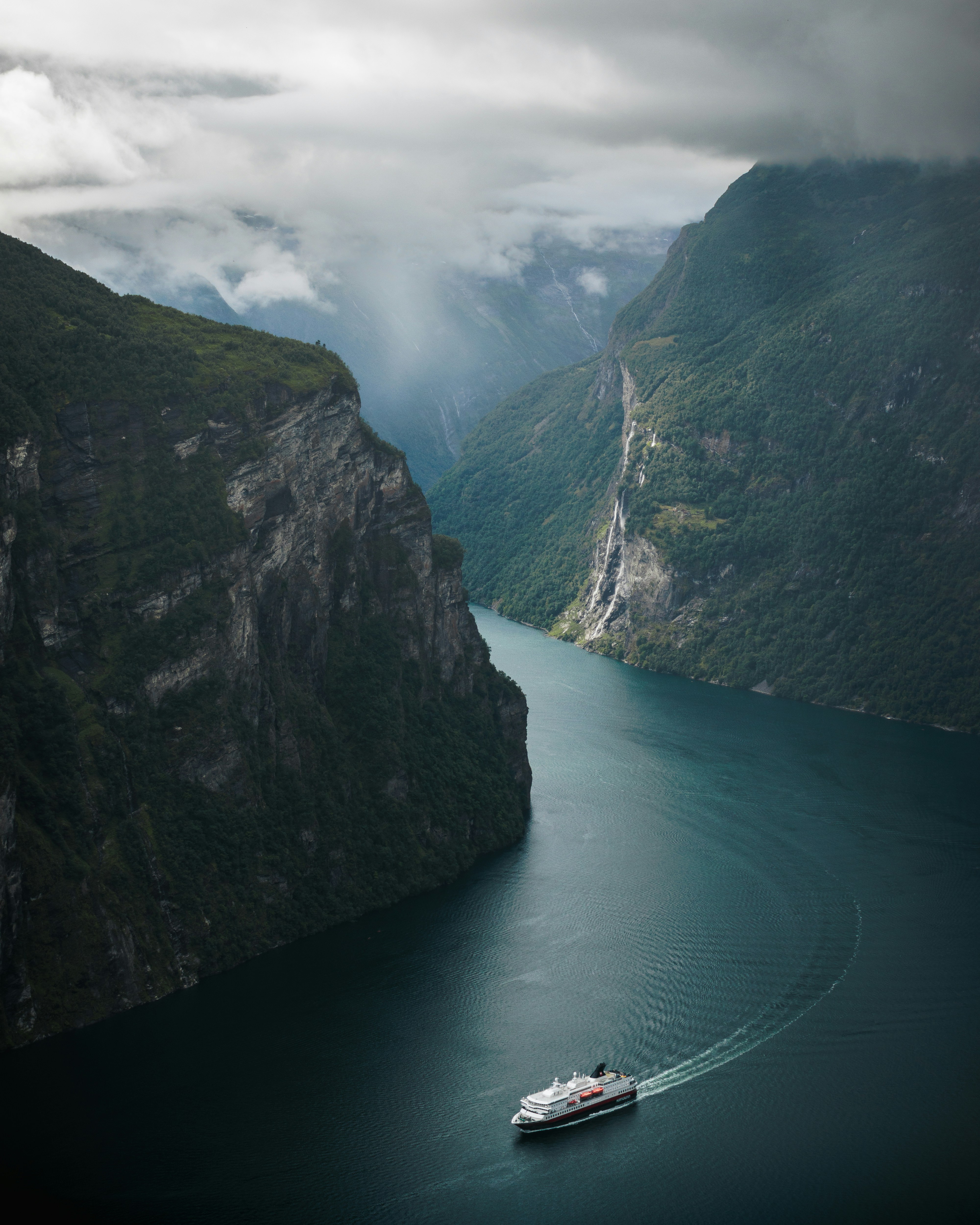 An expedition ship sailing through a deep Norwegian fjord, mountains rising steeply on both sides