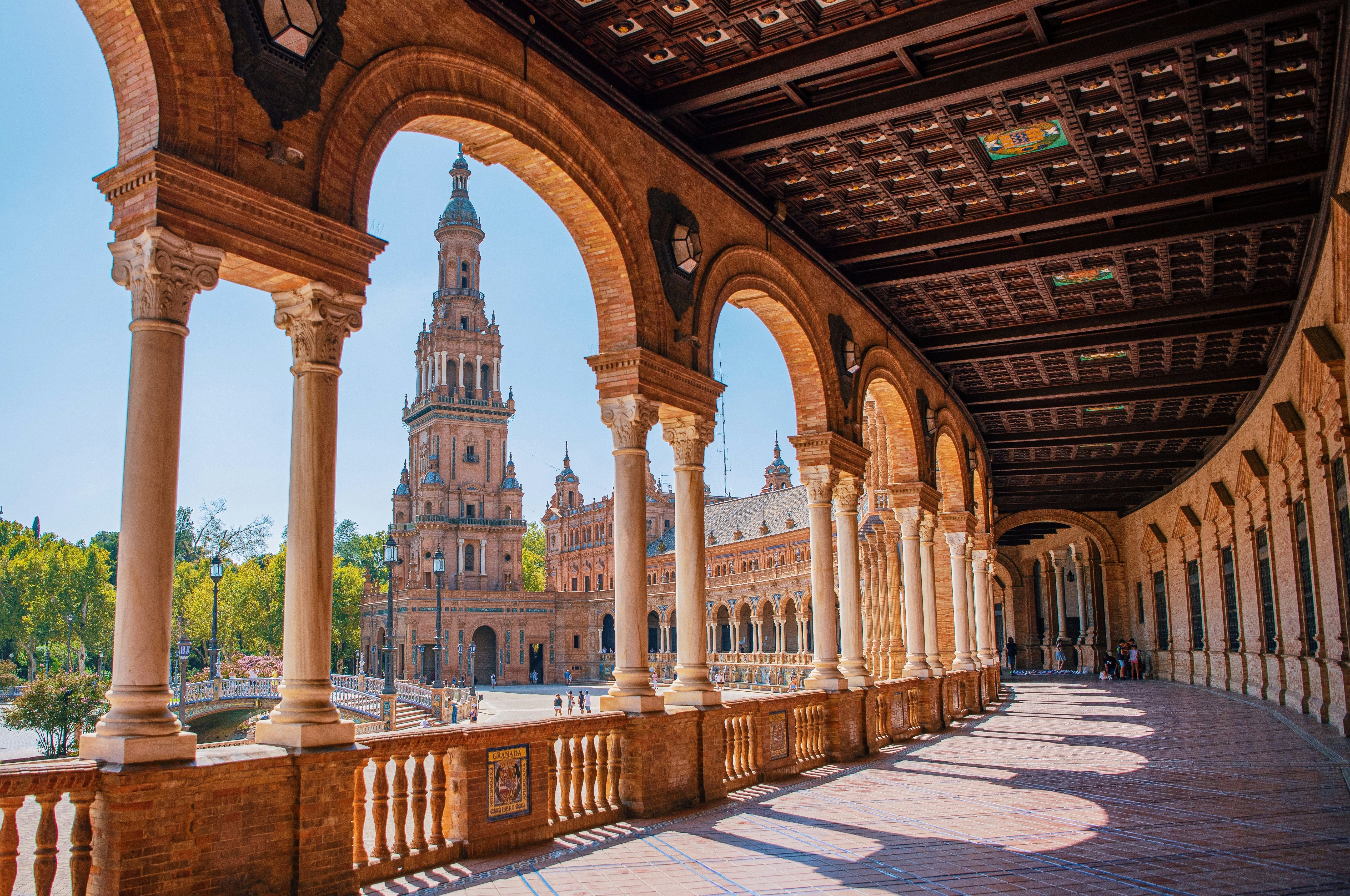 The colonnaded gallery of the Plaza de Espa&ntilde;a in Seville, warm brick arches framing a clear blue sky
